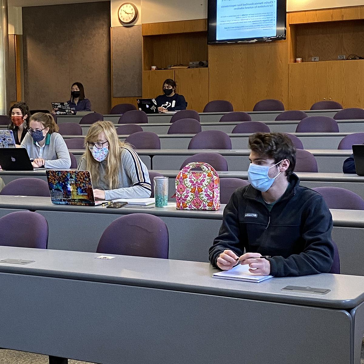 戴着面具的学生们在教室里隔着一段距离坐着, 在笔记本和笔记本电脑上工作